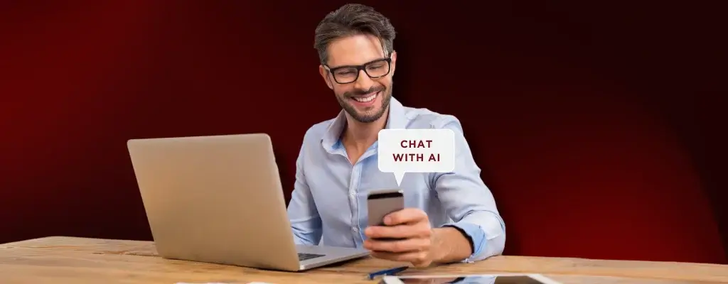 A man with glasses holding his phone while in front of a laptop